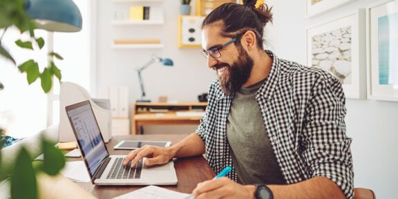 Man sitting at desk smiling, looking at laptop screen, pen in hand