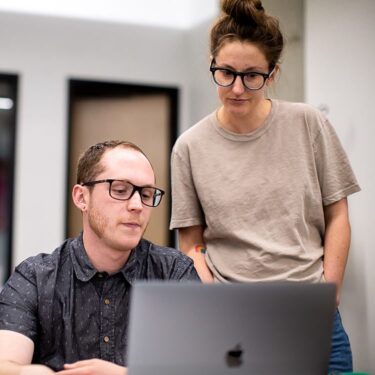 Man working on his laptop while a woman stands over him and watches.