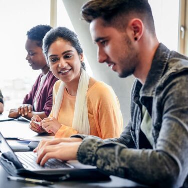 Man working on her laptop while a woman is sitting next to him smiling and watching.