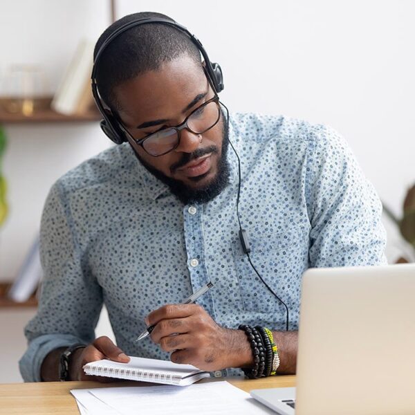 Man wearing headphones writing in his notebook while looking at his laptop.