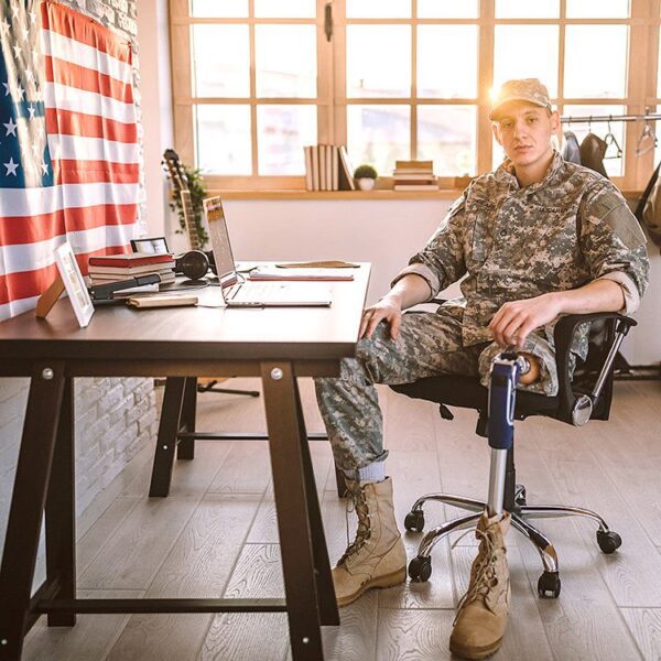 Man in military uniform with a prosthetic leg sitting at a desk in front of his laptop.