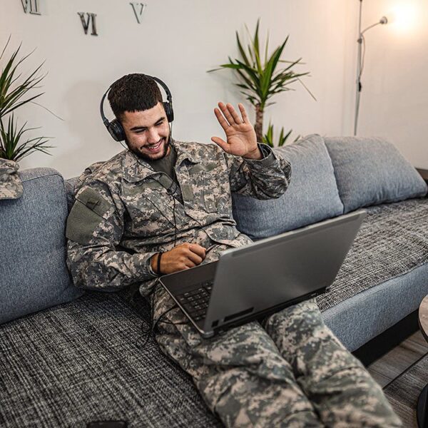 Man in military uniform sitting on a couch on a video call waving to his laptop screen.