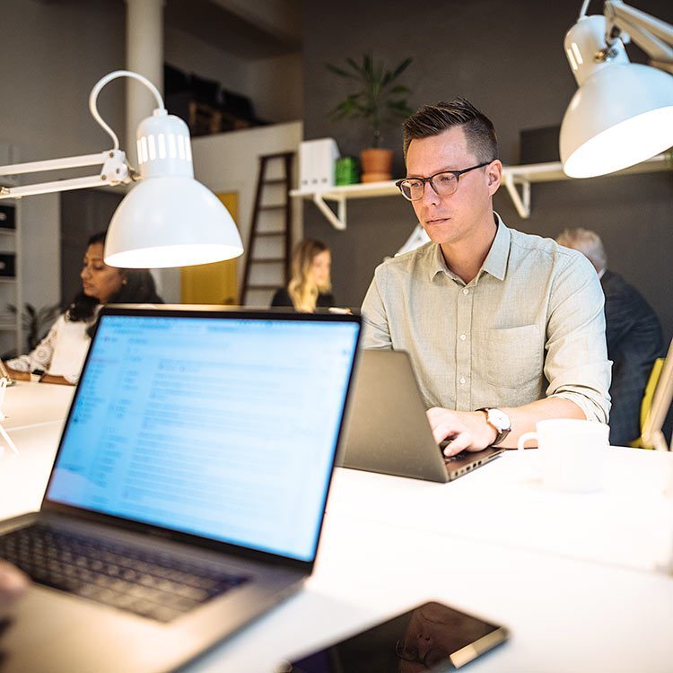 Man working on his laptop in an office surrounded by other people.