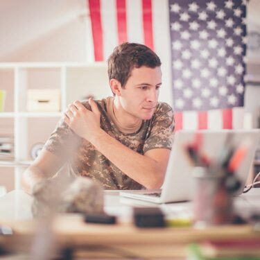 Person in military uniform working on his laptop with an American Flag behind him. Researching VET TEC program funding opportunities.