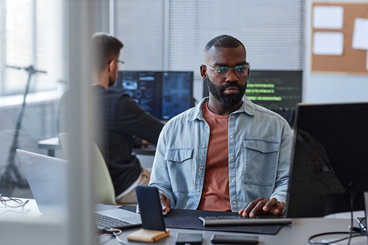 Office professional working at a computer workstation with code displayed on monitors in the background.