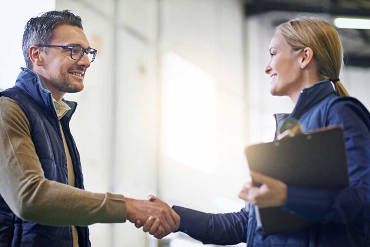 Two professionals smiling and shaking hands during a workplace interaction, with a clipboard visible.
