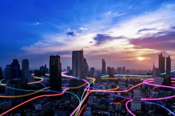 City skyline at sunset with high-rise buildings silhouetted against a colorful sky, overlaid with glowing neon light trails in pink, blue, red, and yellow weaving through the streets to suggest digital connectivity and data flow.